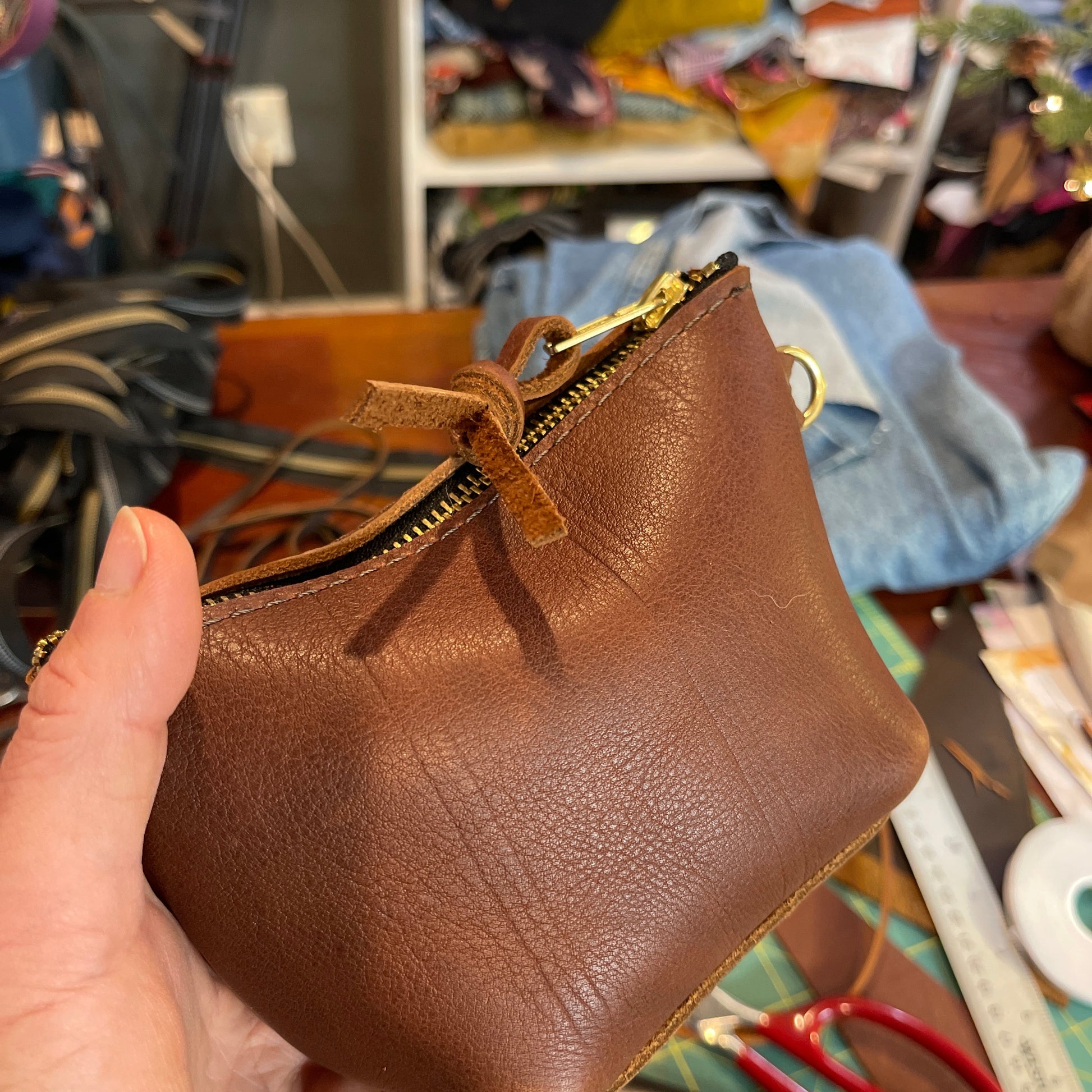 Brown leather pouch with a gold zipper held by a hand in a workshop setting.
